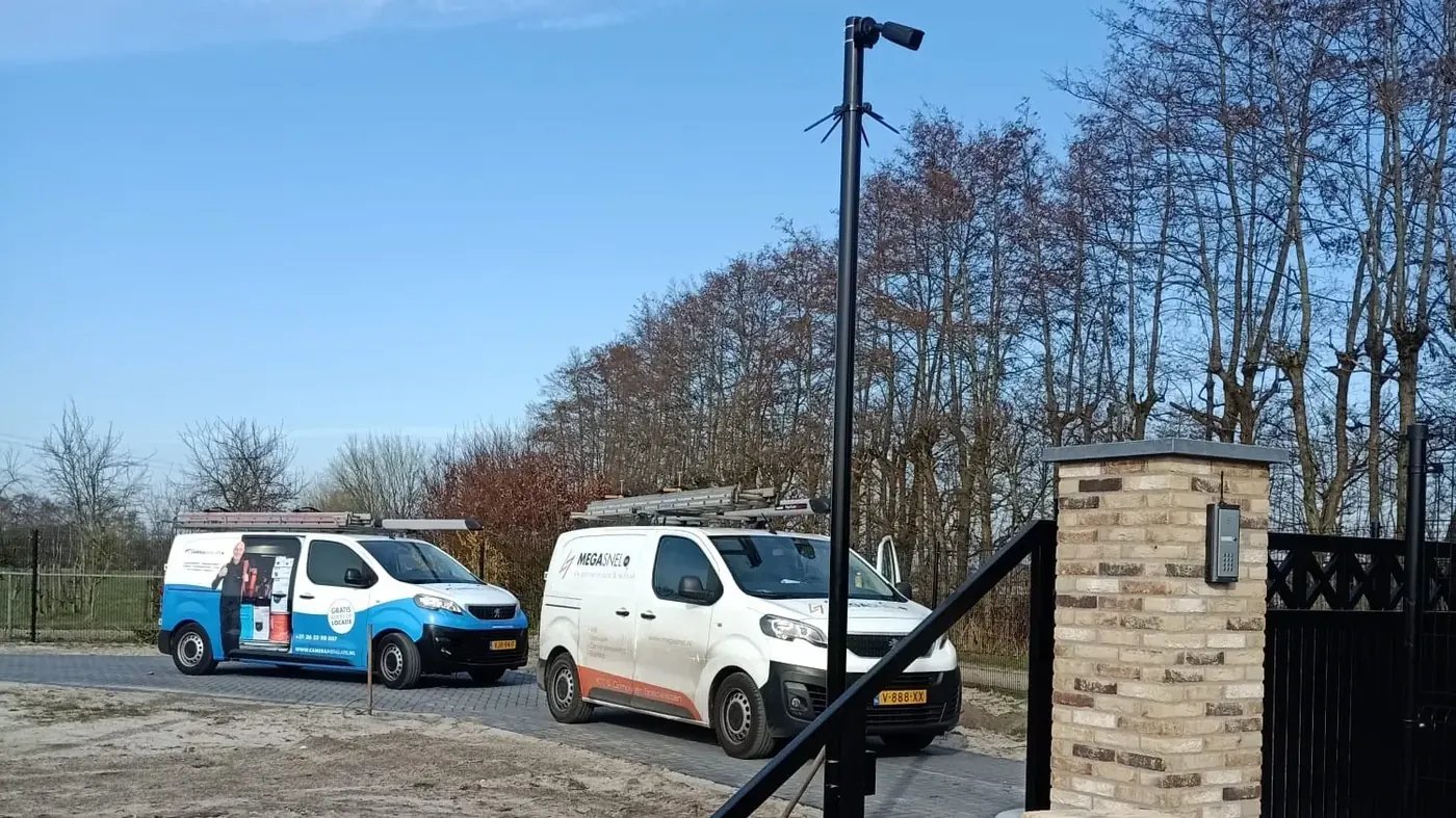 Camerapaal bij de oprit van een woning, met twee servicebussen op de weg