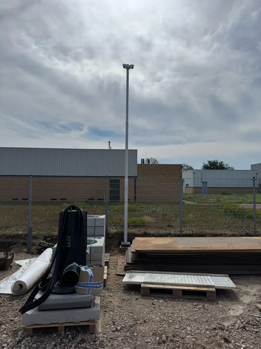 Camerapaal op een bouwterrein, met pallets bouwmateriaal en hekwerk eromheen