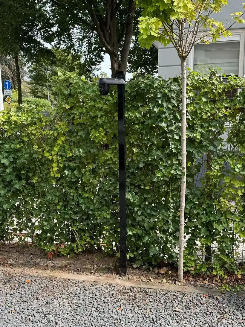 Zwarte camerapaal met camera voor een begroeide wand langs een grindpad